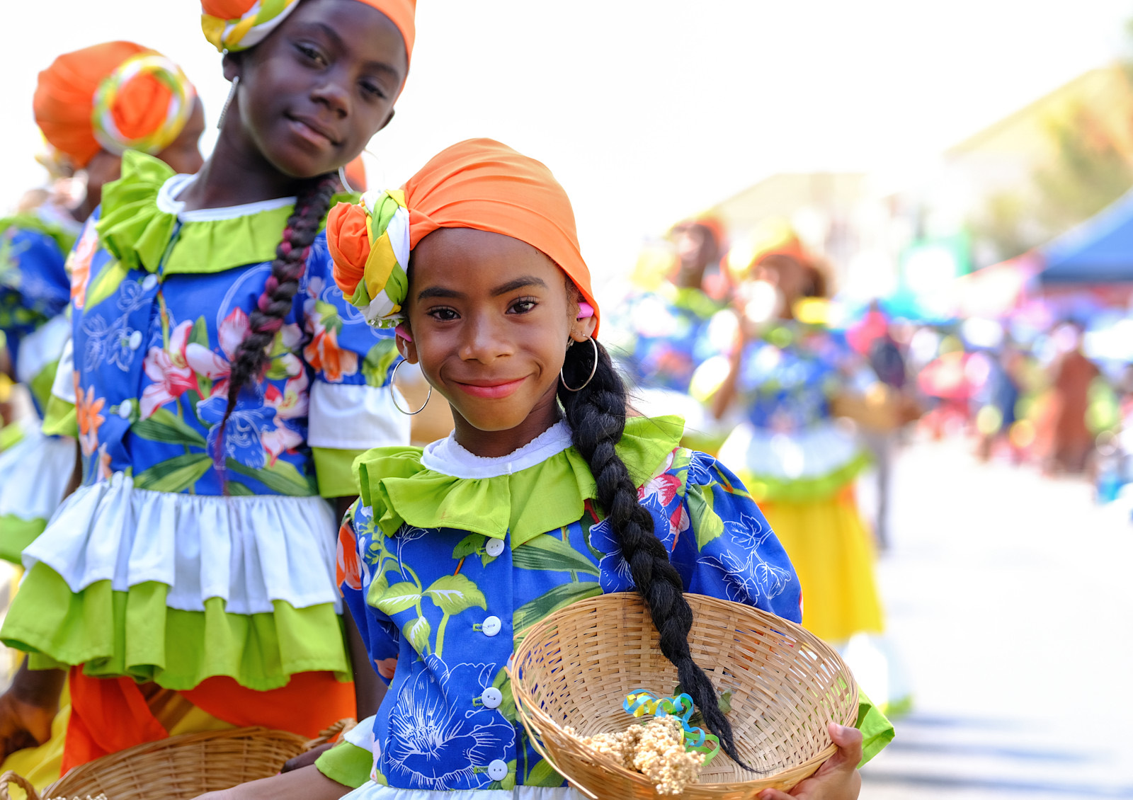 A young girl in a blue floral Seú costume and orange headwrap carrying a woven harvest basket